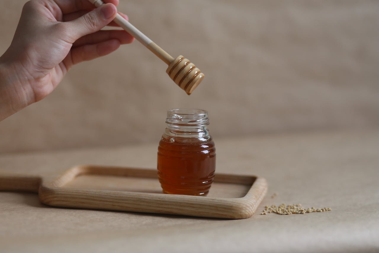 A hand holding a honey dipper over a jar on a wooden tray, showcasing the golden liquid.
